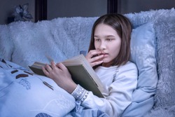 Surprised teenage girl reads an exciting book lays on bed in a cozy bedroom interior. Female college student holding novel and covering mouth with hand. Scary, mystery, suspense tale concepts.