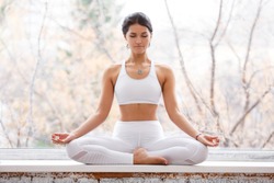 Ð¡harming young woman sitting in lotus pose and meditating with closed eyes during yoga classes on the windowsill in the gym