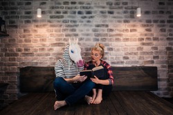 Unusual couple reading a book in stylish apartment. Attentive girl sits on bed with funny man in comical mask. Young woman with strange boyfriend at home.