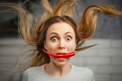 A girl with a red hot pepper in her teeth. Chili for cooking. The spicy seasoning is very stinging in the mouth. My hair stood on end. Bite the pepper and get burned. Emotional woman.
