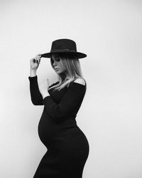 A young beautiful pregnant girl in a black dress on a light background demonstrates her belly. Woman model in a beautiful hat is expecting a baby. Stylish mom