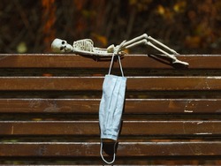 Photograph of a skeleton lies on a wooden bench at night. Tired abstract skeleton toy. He holding surgical mask. Natural dark background. Halloween concept.