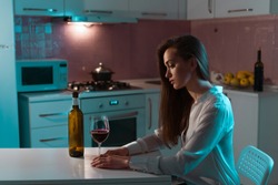 Lonely, unhappy, sad beautiful young woman in a blouse with glass of red wine is drinking alone in evening at home. Female alcoholism and alcohol addiction