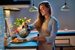 Happy smiling attractive young woman housewife pouring and making tea using electric kettle for evening coffee break at modern loft style kitchen