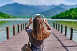 Girl traveler listening to calm music on wireless headphones standing alone on pier with lake and mountains view and enjoying serene quiet peaceful atmosphere 