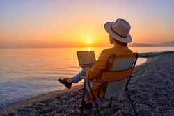 Satisfied free millennial freelancer woman using computer and sitting on beach by calm sea at sunset. Enjoyment of dream office remote work concept