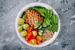 Healthy buddha bowl lunch with grilled chicken, quinoa, spinach, avocado, brussels sprouts, tomatoes, cucumbers  on dark gray background. Top view.