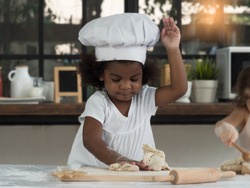 Adorable dark skinned African girl wearing chef hat playing with baking dough in the kitchen at home.