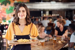 Portrait Of Waitress Serving Food To Customers In Busy Bar Restaurant