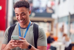 Smiling Male College Student Checking Mobile Phone In Busy Communal Campus Building