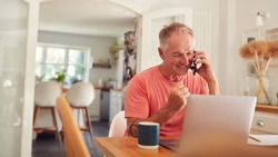 Retired Man On Phone At Home In Kitchen Using Laptop Celebrating Good News Or Winning
