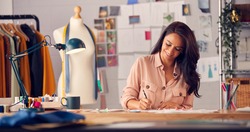 Female Fashion Designer In Studio Working On Sketches And Designs At Desk