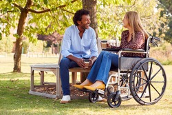 Mature Couple With Woman Sitting In Wheelchair Talking And Drinking Coffee In Park Together