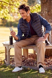 Smiling Man Sitting On Bench In Autumn Park Using Mobile Phone