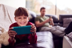 Father Uses Mobile Phone As Son Plays Computer Game On Portable Gaming Device At Home In Pyjamas