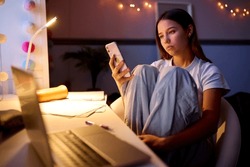 Worried Teenage Girl Sitting At Desk In Bedroom At Home Looking At Mobile Phone At Night