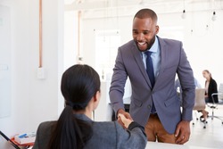 Black businessman and seated woman shaking hands in office