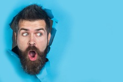 View of male face through hole in blue paper. Surprised bearded man making hole in paper. Copy space for advertising, to insert text or slogan. Discount, sale, season sales.
