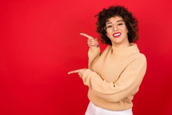 Young beautiful Arab woman wearing knitted sweater standing against red background points aside with  surprised expression with mouth opened, shows something amazing. Advertisement concept.