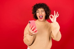 Happy Young beautiful Arab woman wearing knitted sweater standing against red background sending a message on his smartphone or taking a selfie  and making ok sign with his hand.