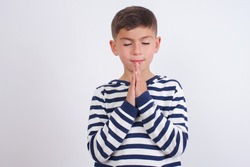 Indoor closeup of Cute Caucasian kid boy wearing knitted sweater against white wall practicing yoga and meditation, holding palms together in namaste, looking calm, relaxed and peaceful.