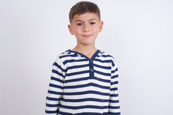 Happy little cute Caucasian boy kid wearing stripped t-shirt against white wall looking at camera with charming cute smile.