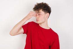 Very upset, young caucasian man with curly hair wearing red T-shirt on ​white background touching nose between closed eyes, wants to cry, having stressful relationship or having troubles with work