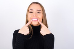 Strong healthy straight white teeth. Close up portrait of happy Young Caucasian girl wearing black knitted over white background with beaming smile pointing on perfect clear white teeth.