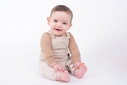 Adorable baby boy wearing beige overalls sitting on white background looking at camera and smiling. 