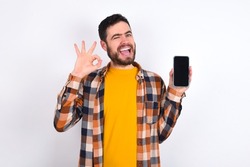 Excited young caucasian man wearing plaid shirt over white background showing smartphone blank screen, blinking eye and doing ok sign with hand.  Advertisement concept.