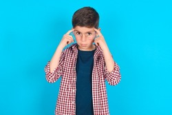 Serious concentrated caucasian kid boy wearing plaid shirt over blue background keeps fingers on temples, tries to ease tension, gather with thoughts and remember important information for exam