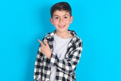 caucasian kid boy wearing plaid shirt over blue background pointing away and smiling to you. Look over there!