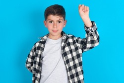 caucasian kid boy wearing plaid shirt over blue background feeling serious, strong and rebellious, raising fist up, protesting or fighting for revolution.