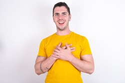Happy smiling Young caucasian man wearing yellow t-shirt over white background has hands on chest near heart. Human emotions, real feelings and facial expression concept.