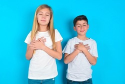 two kids boy and girl standing over blue background closes eyes and keeps hands on chest near heart, expresses sincere emotions, being kind hearted and honest. Body language and real feelings concept.