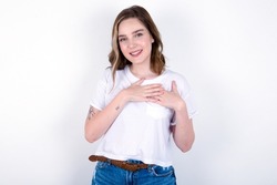 Honest young caucasian woman wearing white T-shirt over white background keeps hands on chest, touched by compliment or makes promise, looks at camera with great pleasure.