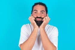 Caucasian man with beard wearing white t-shirt over blue background with surprised expression keeps hands under chin keeps lips folded makes funny grimace