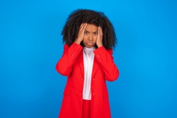 young businesswoman with afro hairstyle wearing red over blue wall holding head in hands with unhappy expression watching sad movie about animals and trying not to cry.
