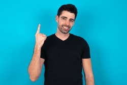 Young caucasian man wearing black T-shirt over blue background smiling and looking friendly, showing number one or first with hand forward, counting down