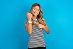 Young beautiful woman wearing striped t-shirt In hurry pointing to wrist watch, impatience, looking at the camera with relaxed expression