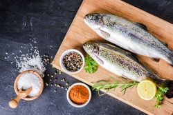 Fresh rainbow trouts on wooden board on dark stone table with salt, pepper, lemon and rosemay ingredients. Tasty fishes preparing for lunch.
