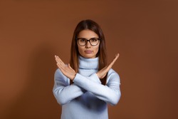 Forbidden. Pretty caucasian woman with dark hair wearing glasses and light blue sweater makes denial hand gesture, keeps arms crossed over chest, demonstrates stop sign, isolated on brown background