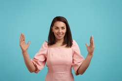 Pleased cheerful smiling european woman with dark hair in pink dress makes big sign with both hands, shapes quite huge object, impressed by size, shapes huge box, isolated on blue background