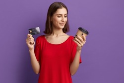 Best coffee in app. Happy young pretty brunette girl in red t-shirt holds disposable cup and showing a debit or credit card. Pays by internet for hot drink. Posing over purple wall