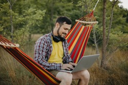 A young man works with his laptop in nature, sitting in a hammock outdoors. Happy man relaxing and working sitting on a hammock in the woods, close-up portrait, wearing shorts and a yellow T-shirt.