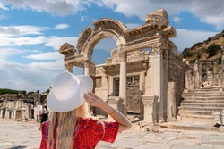 Back view of girl in red dress and white hat watches Temple of Hadrian ruins in antique Ephesus city, Turkey, on a sunny summer day, Efes, Izmir, Turkey