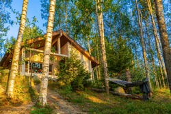 Summer landscape with wooden eco cottage and hammock in forest in evening at sunset. Facade of log cabin among birch trees. Staycation in fresh air, beautiful location, rest in country house.
