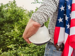 Handsome man holding US Flag and construction helmet against the background of trees, blue sky and sunset. View from the back. Labor and employment concept