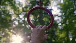 Close up hand of slender athletic woman takes on gymnastic rings. girl in park is doing workout and go in for sport in open area in forest with red gymnastic apparatus at sunset or dawn.