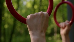 Rack focus close up hand of slender athletic woman takes on gymnastic rings. girl in park is doing workout and go in for sport in open area in forest with red gymnastic apparatus at sunset or dawn.